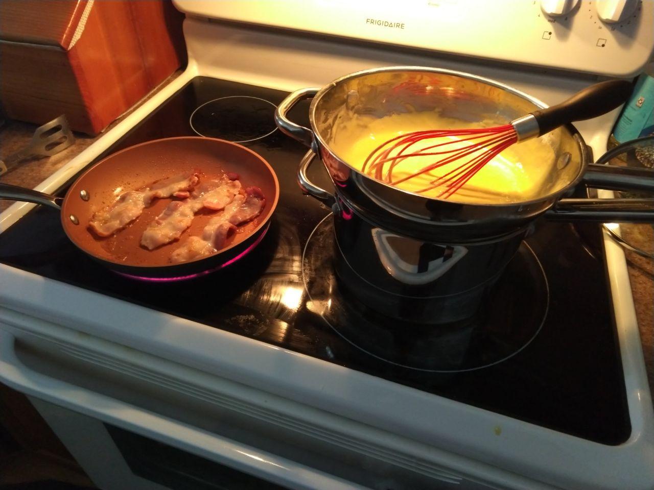 Hollandaise cooking in double boiler with bacon frying in a pan.